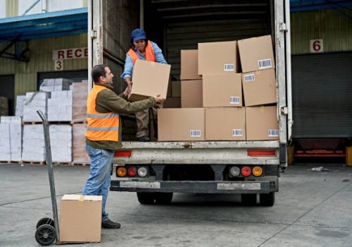 Full length view of late 40s man moving boxes from loading dock with hand truck and lifting into hands of woman organizing merchandise on vehicle.
